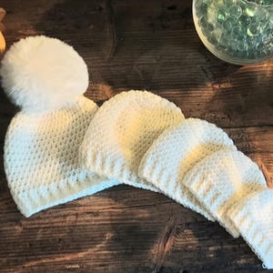May include: Four white crocheted hats with a pom-pom on the largest hat. The hats are arranged in a row from largest to smallest on a wooden surface.