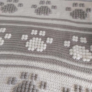 May include: A crocheted blanket with a repeating pattern of paw prints. The blanket is made with white and gray yarn and has a textured, puffy look.