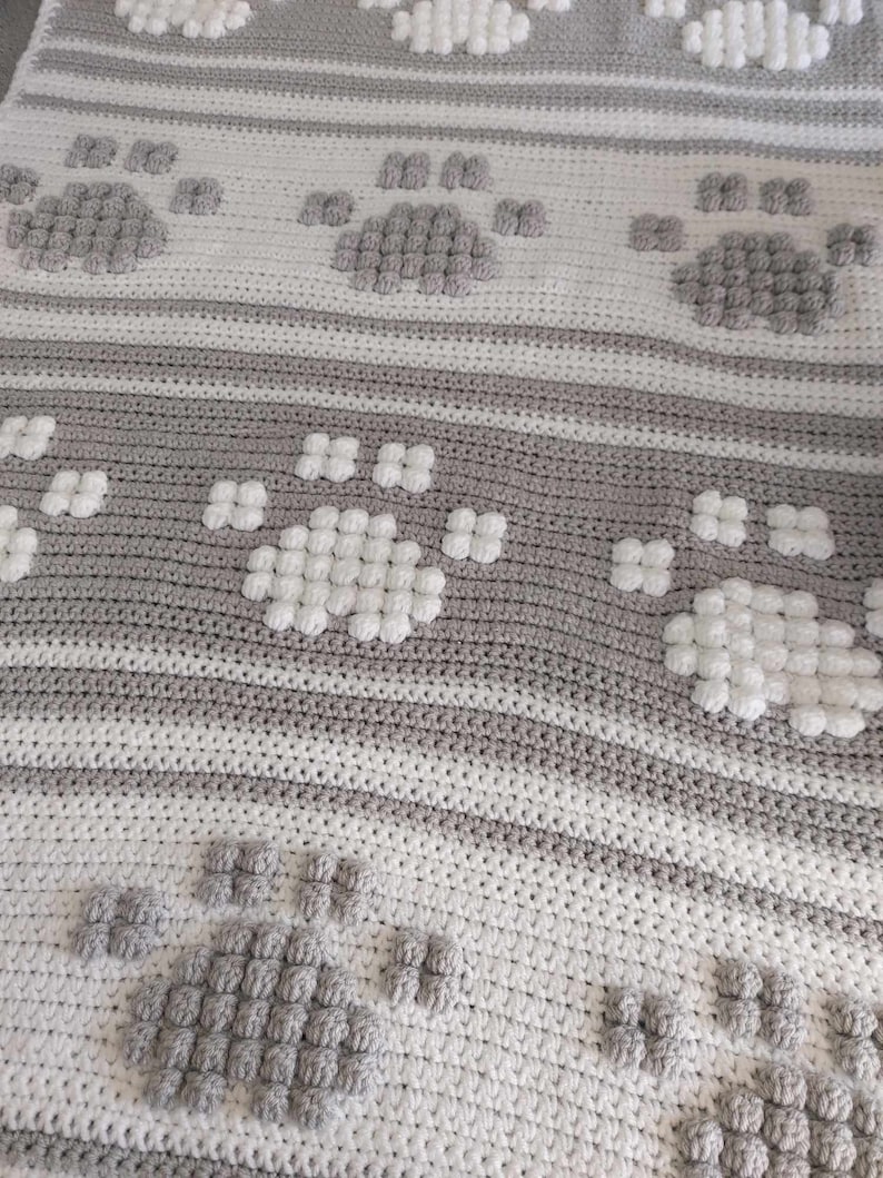 May include: A crocheted blanket with a repeating pattern of paw prints. The blanket is made with white and gray yarn and has a textured, puffy look.