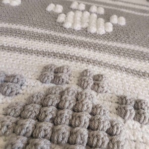 May include: Close-up of a gray and white crocheted blanket with a textured pattern. The blanket features a repeating design of raised, circular stitches.