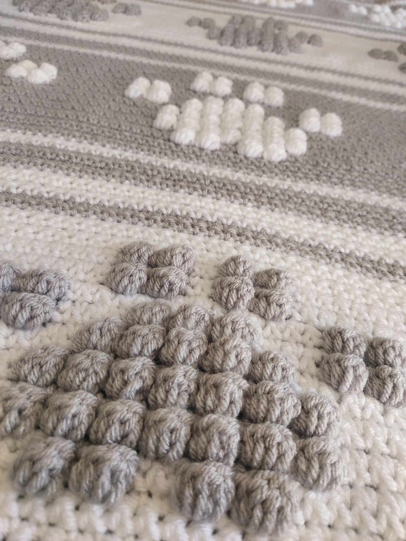 May include: Close-up of a gray and white crocheted blanket with a textured pattern. The blanket features a repeating design of raised, circular stitches.
