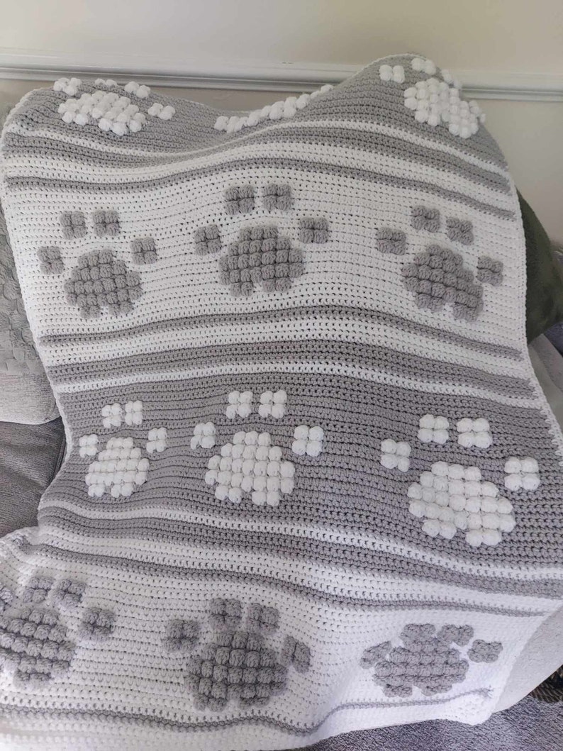 May include: A gray and white crocheted baby blanket with a paw print design. The blanket is made with soft yarn and has a textured surface.