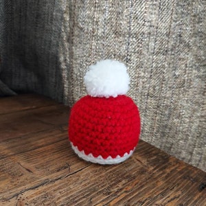 Könnte beinhalten: Eine kleine rot-weiße gehäkelte Weihnachtsmütze mit einem weißen Bommel oben.
