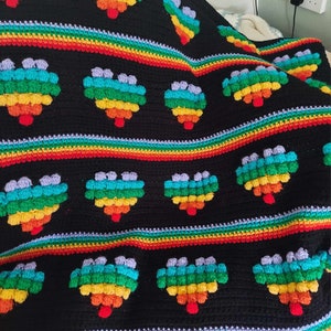 Könnte beinhalten: Eine schwarze Häkeldecke mit regenbogenfarbenen Herzen. Die Herzen bestehen aus einzelnen Maschen in einem Noppenmuster.