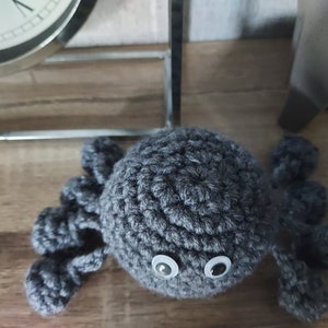 May include: A grey crocheted octopus with white eyes. The octopus has eight tentacles and is sitting on a wooden surface.