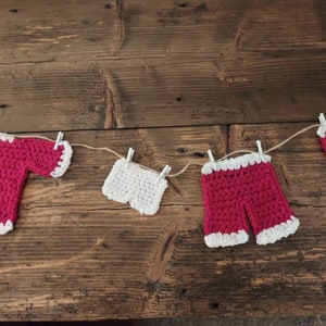 May include: A string of crocheted clothing items, including a red sweater, white underwear, red shorts, and a red hat, are hanging on a clothesline with clothespins.