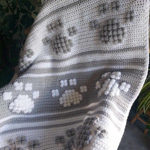 May include: A gray and white crocheted blanket with a paw print design. The blanket is made with a textured yarn and has a soft, cozy feel.