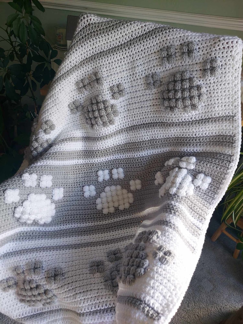 May include: A gray and white crocheted blanket with a paw print design. The blanket is made with a textured yarn and has a soft, cozy feel.