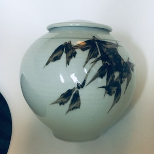 Puede incluir: Un tarro de cerámica blanco con tapa, con un diseño de bambú negro y gris. El tarro tiene una forma redonda y un acabado liso y brillante.