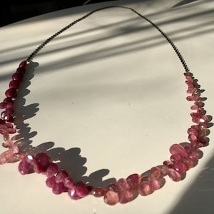 Pode incluir: Um colar longo feito de contas de pedras preciosas rosa e vermelhas. As contas são de forma oval e estão enfiadas em uma corrente prateada.