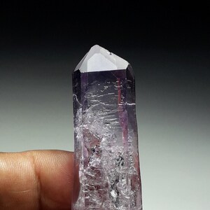 May include: A translucent amethyst crystal with a faceted top. The crystal transitions from a deep purple at the top to a lighter lavender hue. A small, rough stone is attached to the base.
