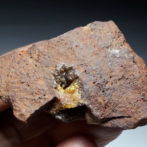May include: A close-up of a rough, brown rock with a yellow and green mineral deposit. The mineral is embedded in the rock and has a crystalline structure.