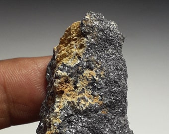 Può includere: Una roccia ruvida, grigia e nera con una piccola macchia di minerale giallo-marrone. La roccia ha un aspetto metallico.