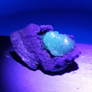 May include: A green crystal cluster embedded in a dark gray rock. The crystal is illuminated with a bright green glow under UV light.