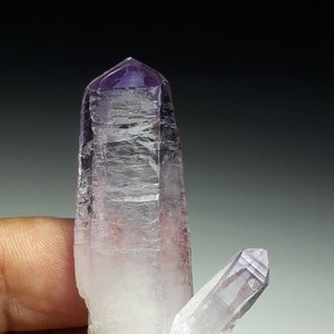 May include: A cluster of two amethyst crystals, one larger and one smaller, with a clear, translucent base and a purple tip. The crystals are pointed and have a natural, rough texture.