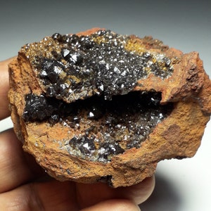 May include: A cluster of dark, shiny crystals embedded in a rough, orange-brown rock. The crystals have a sharp, geometric shape and are arranged in a dense, compact formation.