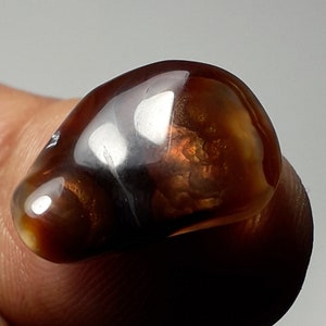 Puede incluir: Una piedra de ágata lisa y pulida con un patrón de color marrón, naranja y blanco. La piedra tiene forma ovalada y un acabado brillante.