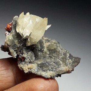 May include: A mineral specimen featuring a cluster of pale yellow crystals. These crystals sit on a matrix of grey and brown minerals. Small, reddish-orange mineral deposits are also visible, creating a striking contrast.