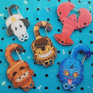 Peut inclure: Plusieurs porte-clés colorés de style dessin animé. Les motifs incluent un chien, un homard et des chats. Chaque porte-clés est doté d'un fermoir argenté et est présenté sur un fond bleu parsemé de points noirs.