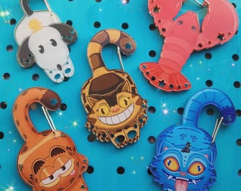 May include: A collection of colorful, cartoon-style keychains featuring various designs. The keychains include a dog, a lobster, a cat, a blue cat, and an orange cat. Each keychain has a silver clasp and is set against a blue background.