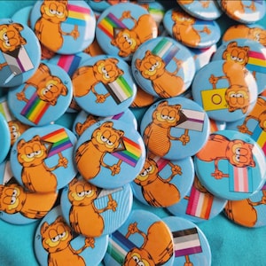 Peut inclure: Une collection de badges boutonnés colorés représentant le personnage de dessin animé Garfield tenant différents drapeaux de fierté, notamment les drapeaux arc-en-ciel, bisexuel, transgenre et ace. Les boutons sont sur une surface bleue.