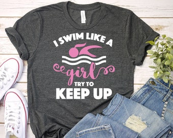 Puede incluir: Una camiseta gris con el texto "I swim like a girl try to keep up" en blanco. El texto "girl" está en rosa. Una silueta de nadador también está en rosa.