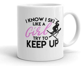Puede incluir: Taza de café de cerámica blanca con un gráfico en blanco y negro de un esquiador y el texto "I know I ski like a girl try to keep up". La palabra "girl" está en rosa.