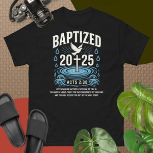 Op de afbeelding: Zwart T-shirt met een witte duif en kruis grafische afbeelding. De tekst "BAPTIZED 20+25" staat boven de afbeelding. De tekst "ACTS 2:38" staat onder de afbeelding. De tekst "REPENT AND BE BAPTIZED, EVERY ONE OF YOU, IN THE NAME OF JESUS CHRIST FOR THE FORGIVENESS OF YOUR SINS. AND YOU WILL RECEIVE THE GIFT OF THE HOLY SPIRIT." staat onder de tekst "ACTS 2:38".
