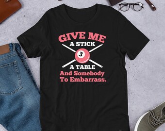 Puede incluir: Camiseta negra con un gráfico blanco que dice "Give me a stick, a table and somebody to embarrass." El gráfico también incluye una bola de billar roja con el número 3 y dos tacos de billar cruzados.