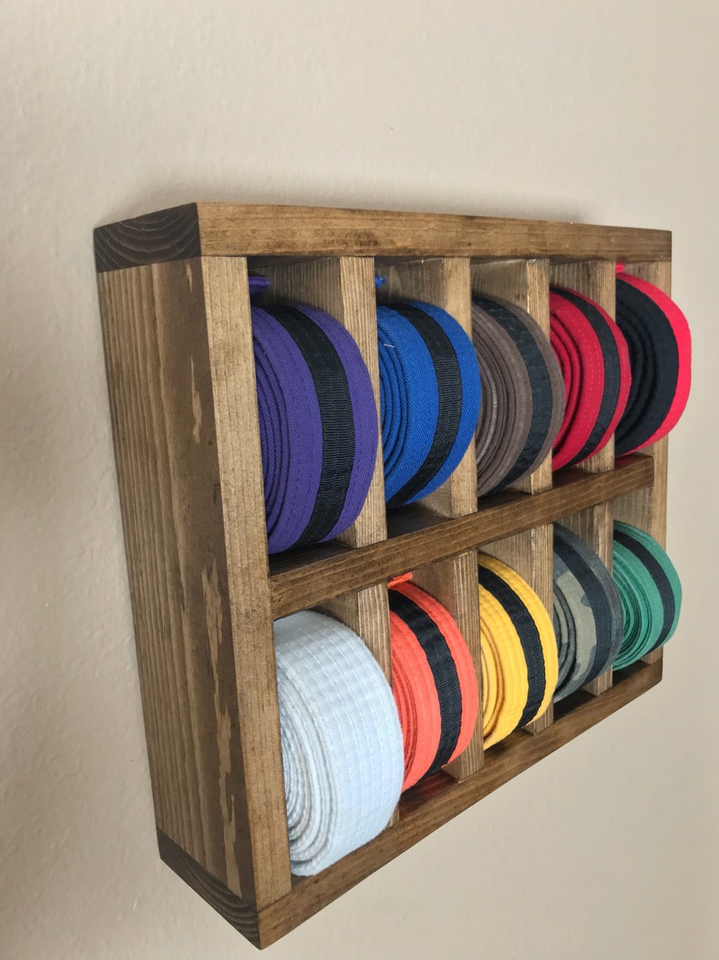 Puede incluir: Estante de pared de madera con 10 compartimentos para guardar cinturones de artes marciales. El estante est&aacute; te&ntilde;ido de un color marr&oacute;n oscuro y tiene un aspecto r&uacute;stico. Los cinturones est&aacute;n dispuestos en los compartimentos, con los colores visibles.