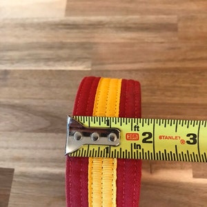 Puede incluir: Una cinta de tela a rayas rojas y amarillas medida con una cinta m&eacute;trica amarilla que dice "25ft" y "Stanley". La cinta tiene 2 pulgadas de ancho.