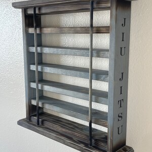 May include: A gray wooden wall shelf with five shelves and black metal supports. The word "JIU JITSU" is engraved on the right side of the shelf.