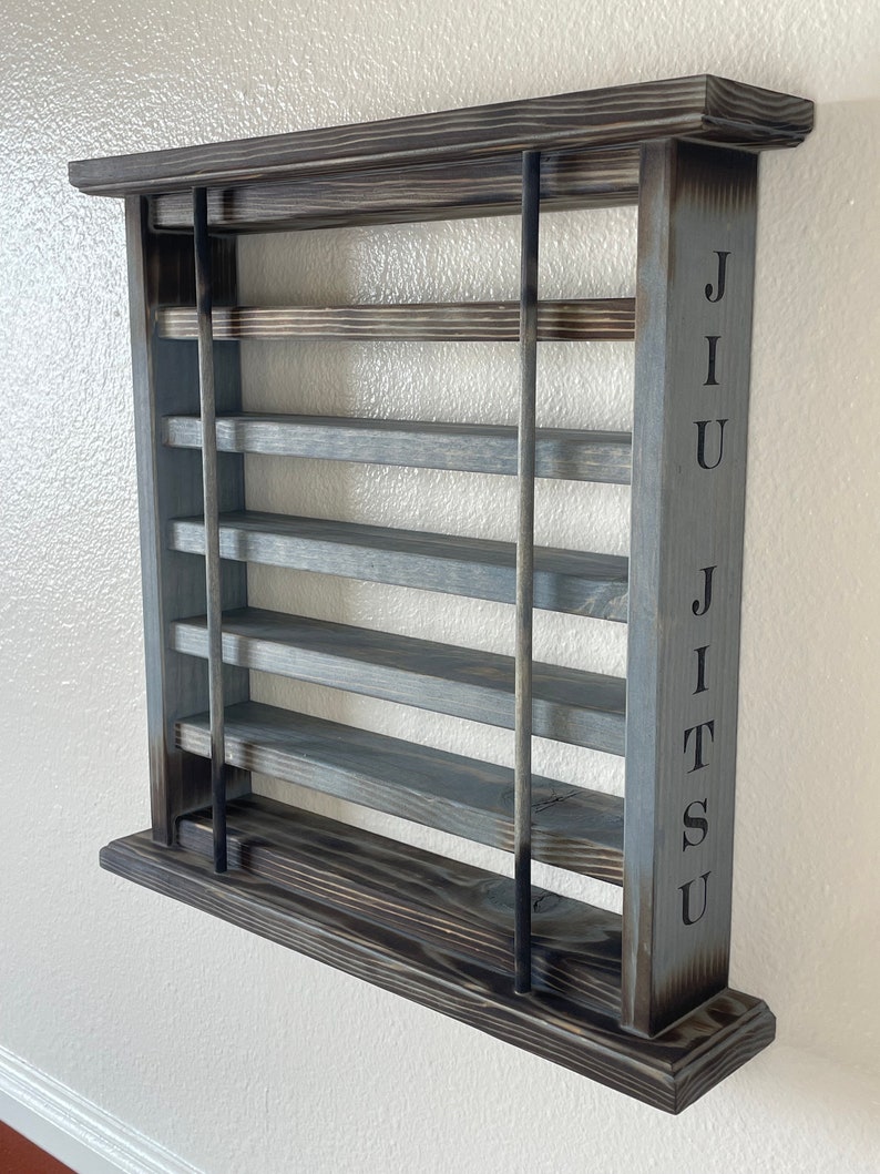 May include: A gray wooden wall shelf with five shelves and black metal supports. The word "JIU JITSU" is engraved on the right side of the shelf.