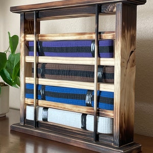 May include: A wooden belt rack with four shelves for storing martial arts belts. The rack is made of dark wood with a distressed finish. The shelves are made of light wood and have black metal rods to hold the belts in place. The rack is sitting on a wooden surface.