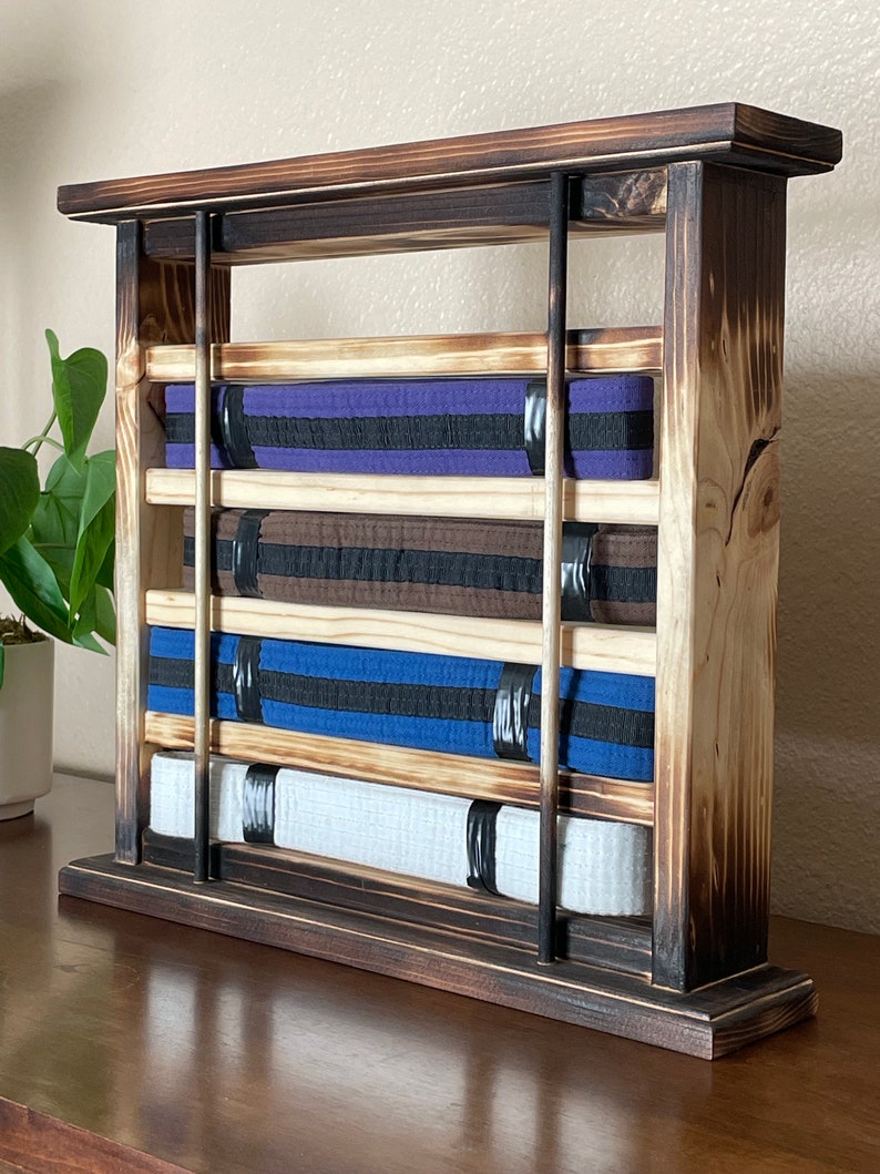 May include: A wooden belt rack with four shelves for storing martial arts belts. The rack is made of dark wood with a distressed finish. The shelves are made of light wood and have black metal rods to hold the belts in place. The rack is sitting on a wooden surface.