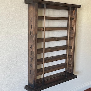 May include: A dark brown wooden wall-mounted shelf with the word "JIU JITSU" engraved on the side. The shelf has six shelves and is designed to hold belts.