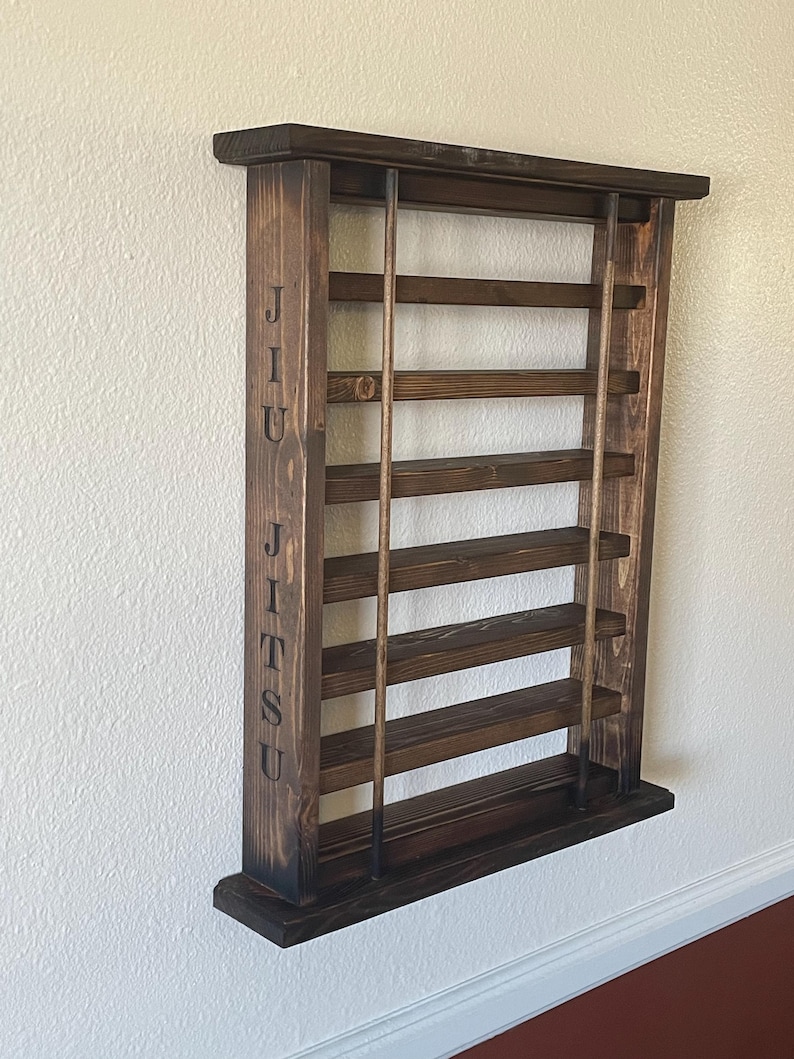 May include: A dark brown wooden wall-mounted shelf with the word "JIU JITSU" engraved on the side. The shelf has six shelves and is designed to hold belts.