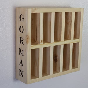 Puede incluir: Una estanter&iacute;a de pared de madera con nueve compartimentos, cada uno con una abertura rectangular. La estanter&iacute;a est&aacute; pintada en un color madera claro y tiene el nombre "GORMAN" escrito verticalmente en el lado izquierdo.