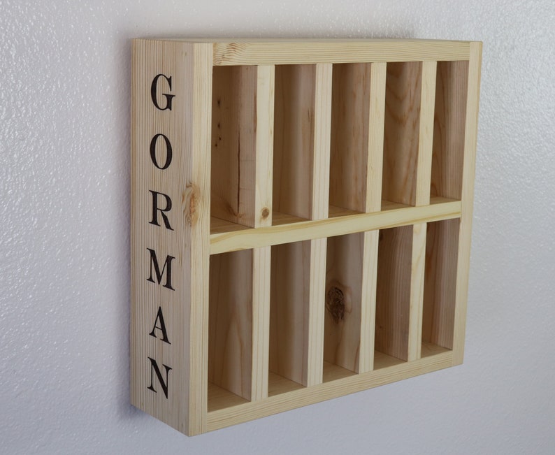 Puede incluir: Una estanter&iacute;a de pared de madera con nueve compartimentos, cada uno con una abertura rectangular. La estanter&iacute;a est&aacute; pintada en un color madera claro y tiene el nombre "GORMAN" escrito verticalmente en el lado izquierdo.