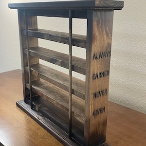 May include: A wooden shelf unit with four shelves and a base. The shelf unit is stained dark brown and has a rustic look. The side of the shelf unit has the words "ALWAYS EARNED NEVER GIVEN" engraved into the wood.