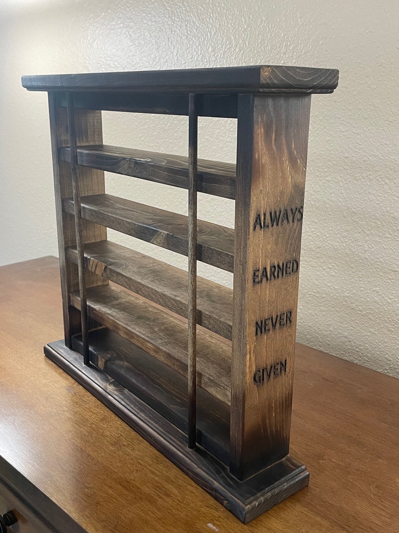 May include: A wooden shelf unit with four shelves and a base. The shelf unit is stained dark brown and has a rustic look. The side of the shelf unit has the words "ALWAYS EARNED NEVER GIVEN" engraved into the wood.
