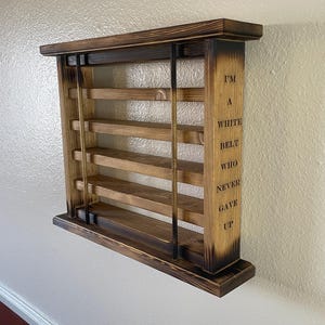 May include: A wooden wall shelf with five shelves and a burnt wood finish. The shelf has a quote on the side that reads "I'm a white belt who never gave up".