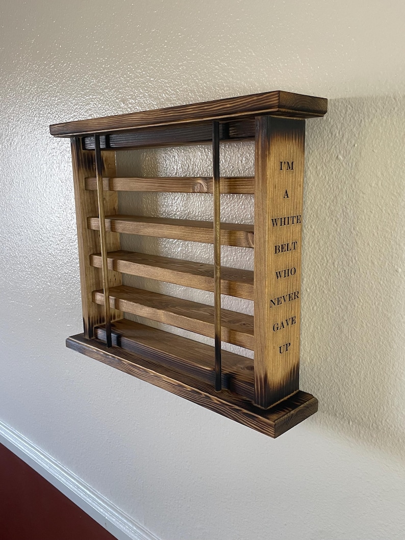 May include: A wooden wall shelf with five shelves and a burnt wood finish. The shelf has a quote on the side that reads "I'm a white belt who never gave up".
