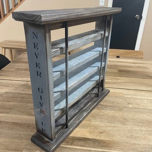 May include: A wooden shelf unit with five shelves, stained in a dark brown color with a gray wash. The shelf unit has a sign on the side that reads "NEVER GIVE UP".