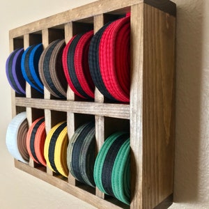 Puede incluir: Un estante de pared de madera con 12 ranuras para guardar cinturones de artes marciales. El estante est&aacute; te&ntilde;ido de un color marr&oacute;n oscuro y tiene un aspecto r&uacute;stico. Los cinturones est&aacute;n ordenados por color, comenzando por el blanco y terminando por el verde.