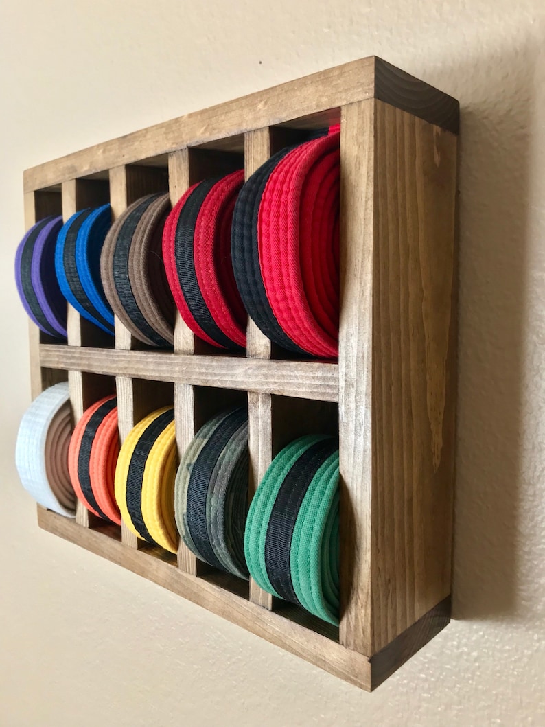 Puede incluir: Un estante de pared de madera con 12 ranuras para guardar cinturones de artes marciales. El estante est&aacute; te&ntilde;ido de un color marr&oacute;n oscuro y tiene un aspecto r&uacute;stico. Los cinturones est&aacute;n ordenados por color, comenzando por el blanco y terminando por el verde.