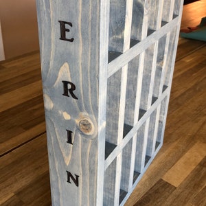 Puede incluir: Una caja de madera gris con una cuadr&iacute;cula de compartimentos. La palabra "ERIN" est&aacute; escrita verticalmente en el lateral de la caja.