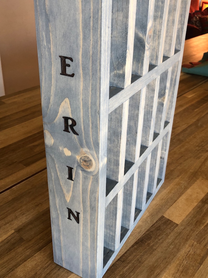 Puede incluir: Una caja de madera gris con una cuadr&iacute;cula de compartimentos. La palabra "ERIN" est&aacute; escrita verticalmente en el lateral de la caja.
