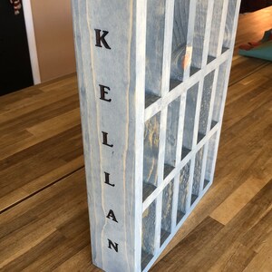 Puede incluir: Una caja de madera gris con una cuadr&iacute;cula de compartimentos. La caja est&aacute; etiquetada "KELLAN" en letras negras.