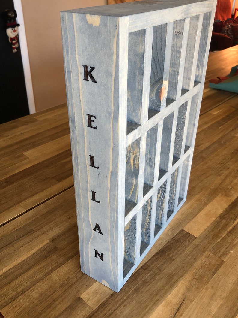 Puede incluir: Una caja de madera gris con una cuadr&iacute;cula de compartimentos. La caja est&aacute; etiquetada "KELLAN" en letras negras.